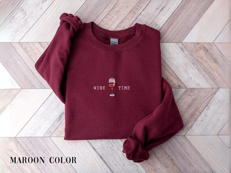May include: Maroon-colored sweatshirt with the words "WINE TIME" and a wine glass graphic. The sweatshirt is displayed on a light wood-grain surface. The sleeves are folded, and the words "MAROON COLOR" are at the bottom.