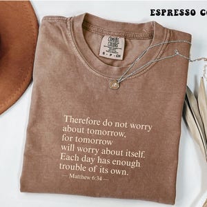 May include: Espresso-colored t-shirt with the text "Therefore do not worry about tomorrow, for tomorrow will worry about itself. Each day has enough trouble of its own. Matthew 6:34." A silver necklace with a pendant rests on the shirt.