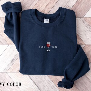 May include: Navy blue sweatshirt with the words "WINE TIME" and a graphic of a wine glass. The sweatshirt is a solid color and has a crew neck and long sleeves. The text and graphic are in white and red.