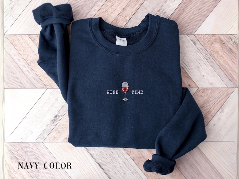 May include: Navy blue sweatshirt with the words "WINE TIME" and a graphic of a wine glass. The sweatshirt is a solid color and has a crew neck and long sleeves. The text and graphic are in white and red.