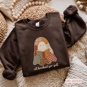 Puede incluir: Una sudadera de cuello redondo en color marrón chocolate oscuro con un gráfico central de un fantasma hecho de varios patrones de tela de patchwork, incluyendo cuadros y flores. El fantasma sostiene un libro abierto con el texto all booked for fall.