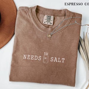 May include: Espresso-colored t-shirt with the text "NEEDS SALT" and a salt shaker graphic. A silver necklace with a pendant rests on the shirt. A brown hat and dried plant are in the background.