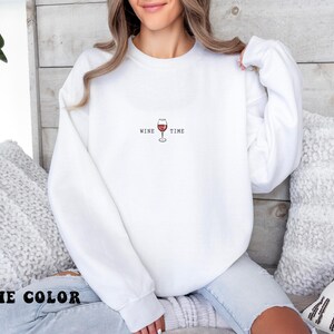 May include: White sweatshirt with the text "WINE TIME" and a graphic of a wine glass in red. The sweatshirt is a crew neck style. The image shows the sweatshirt on a person.