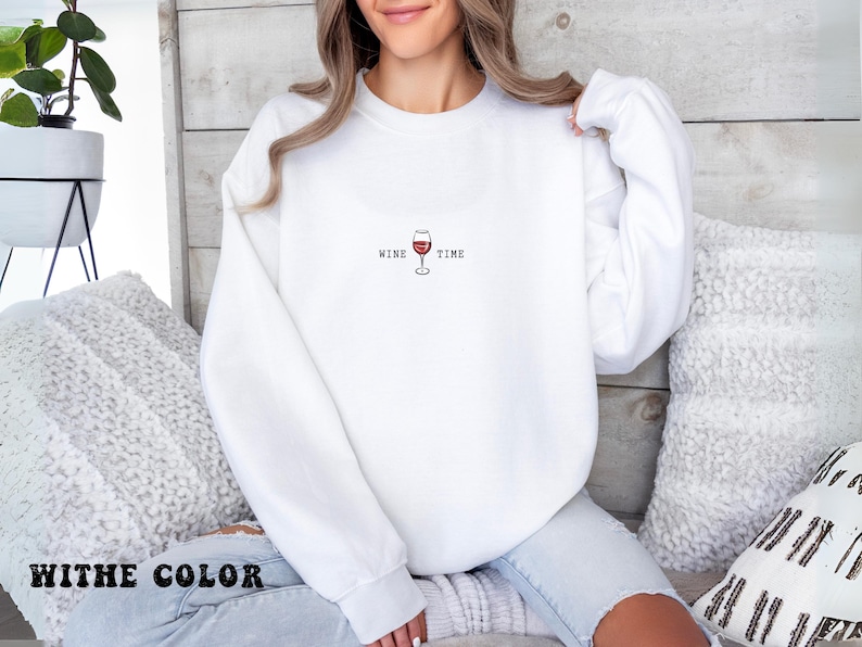 May include: White sweatshirt with the text "WINE TIME" and a graphic of a wine glass in red. The sweatshirt is a crew neck style. The image shows the sweatshirt on a person.