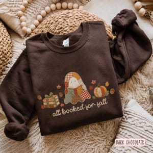 Puede incluir: Una sudadera de cuello redondo en color marrón oscuro con un gráfico de un fantasma leyendo un libro, una pila de libros y una calabaza, todo con un patrón de patchwork. El texto "all booked for fall" aparece en color crema bajo el gráfico.