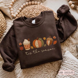 Puede incluir: Una sudadera de cuello redondo en color marrón chocolate oscuro con un diseño bordado de una taza de café, una calabaza estampada y una bellota, rodeados de pequeñas hojas de otoño. El texto 'tis the season' está bordado en cursiva debajo.