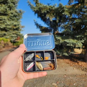 May include: A small, open, rectangular metal tin filled with fishing lures. The lures are in various colors, including silver, orange, pink, and brown. The tin is held in a hand against a backdrop of trees and a blue sky.
