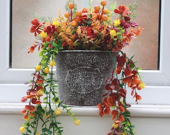 May include: A decorative metal bucket filled with colorful artificial flowers and foliage. The arrangement includes red, orange, and yellow blooms, with trailing green and yellow vines. The bucket sits on a windowsill.