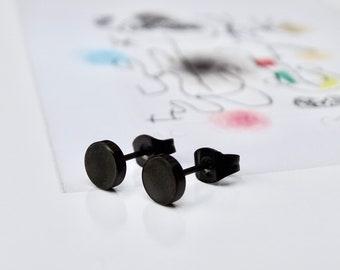 May include: Two black stud earrings with a circular design. The earrings are made of a dark, matte material and are displayed on a white surface with a faint, abstract drawing in the background. The earrings are simple and modern.