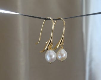 May include: A pair of gold-toned earrings featuring a curved, petal-like metal structure that suspends a single, teardrop-shaped white pearl. The earrings are designed with a long, slender hook for insertion, showcasing a polished, reflective finish.