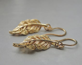 May include: A pair of gold-colored feather earrings. The earrings feature detailed feather designs with a textured surface. The hooks are a simple, curved design. The earrings are displayed against a neutral gray background.