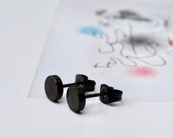 May include: A pair of black stud earrings with a round, flat design. The earrings are made of metal and have a post and butterfly backing.