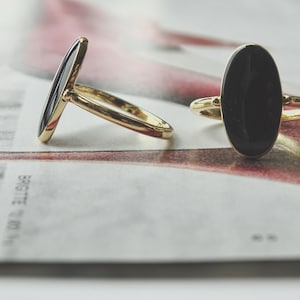 May include: Two gold rings featuring smooth, oval-shaped black stones set in polished metal bands. One ring is positioned upright, displaying the front of the black oval, while the other is tilted to the side, showing the profile of the band.