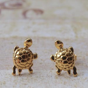 May include: A pair of gold-toned stud earrings shaped like small turtles. Each earring features a detailed, raised shell pattern with a smooth, polished finish. The turtle design includes a small head, four flippers, and a rounded shell, designed to be worn as post-style earrings.