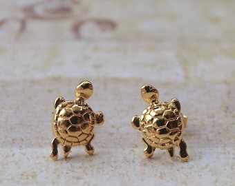 May include: A pair of gold-toned stud earrings shaped like small turtles. Each earring features a detailed, raised shell pattern with a smooth, polished finish. The turtle design includes a small head, four flippers, and a rounded shell, designed to be worn as post-style earrings.