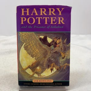 May include: A hardcover book titled Harry Potter and the Prisoner of Azkaban by J.K. Rowling. The purple cover features a yellow moon and an illustration of a person riding a large, winged creature. The text includes the author's name.