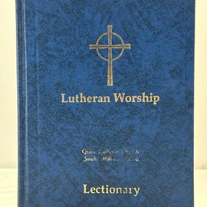 May include: A blue book titled "Lutheran Worship" with gold lettering and a gold cross emblem. The book also has the word "Lectionary" in gold lettering. The cover has a textured pattern and a red ribbon bookmark.