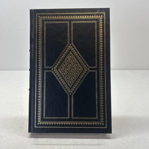 May include: A hardcover book with a dark blue cover and gold decorative accents. The cover features a diamond-shaped design in the center, with rectangular and square gold frames. The edges of the cover are also trimmed in gold.