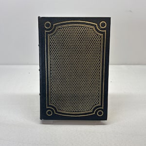 May include: A small, black book with a gold-colored, diamond-patterned center and gold trim. The book has a decorative gold border and gold circles at each corner. The spine of the book is visible on the left side.