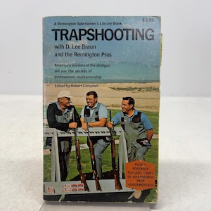 May include: A paperback book titled TRAPSHOOTING with D. Lee Braun and the Remington Pros. The cover features three individuals in shooting vests standing behind a rack of shotguns in a field. Text includes A Remington Sportsman's Library Book.