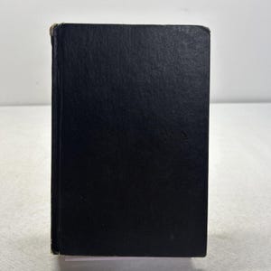 May include: A solid black hardcover book standing upright. The front cover is plain and featureless with a matte texture. The spine edge shows slight wear and fraying at the top corner, revealing a light-colored binding material underneath.