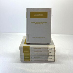 May include: A stack of books titled "The Chimpanzee" with a light gray cover and gold lettering. The top book displays the title and subtitle "Anatomy, Behavior, and Diseases of Chimpanzees". The books are stacked on a white surface.