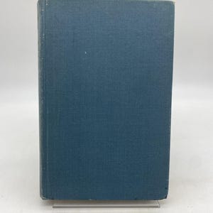 May include: A hardcover book with a teal blue cover. The book is standing upright on a clear plastic stand. The cover is plain and has a slightly textured appearance.