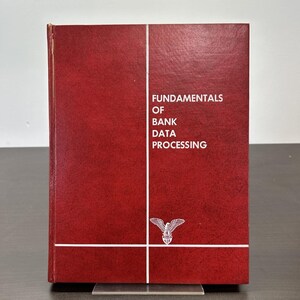 May include: A hardcover book with a textured, burgundy cover. The title "FUNDAMENTALS OF BANK DATA PROCESSING" is printed in white. A white eagle emblem is at the bottom. The book is displayed on a clear stand.