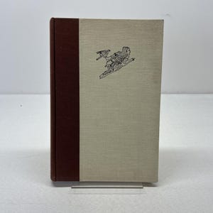 May include: A hardcover book with a brown spine and a light tan cover. The cover features a black illustration of a person with a bird. The book is standing upright on a clear stand.