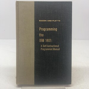 May include: A hardcover book titled Programming the IBM 1401: A Self-Instructional Programmed Manual by Saxon and Plette. The cover features a dark gray fabric texture on the right and a light tan fabric spine on the left with gold text.