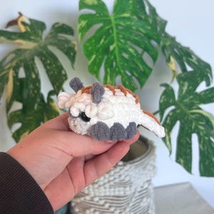 Op de afbeelding: Een kleine, gehaakte Appa-knuffel, met een witte body, grijze voeten en hoorns en bruine accenten. Het handgemaakte speelgoed heeft zwarte ogen en wordt voor een groene plant gehouden.