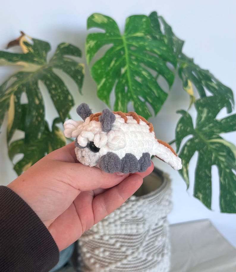 Op de afbeelding: Een kleine, gehaakte Appa-knuffel, met een witte body, grijze voeten en hoorns en bruine accenten. Het handgemaakte speelgoed heeft zwarte ogen en wordt voor een groene plant gehouden.