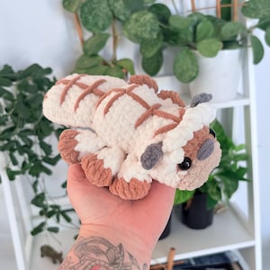 Puede incluir: Un peluche de Appa hecho a mano, de Avatar: La leyenda de Aang. El juguete de ganchillo es blanco con marcas y detalles marrones, incluyendo los pies, las orejas y patrones en forma de cruz en la espalda. El juguete tiene ojos negros y cuernos grises.