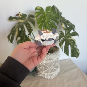 Op de afbeelding: Een kleine, handgemaakte gehaakte figuur van een wezen met grijze, witte en bruine details. De figuur heeft kleine oren en een rond lichaam. De figuur wordt vastgehouden voor een plant met grote groene bladeren met witte en gele vlekken.