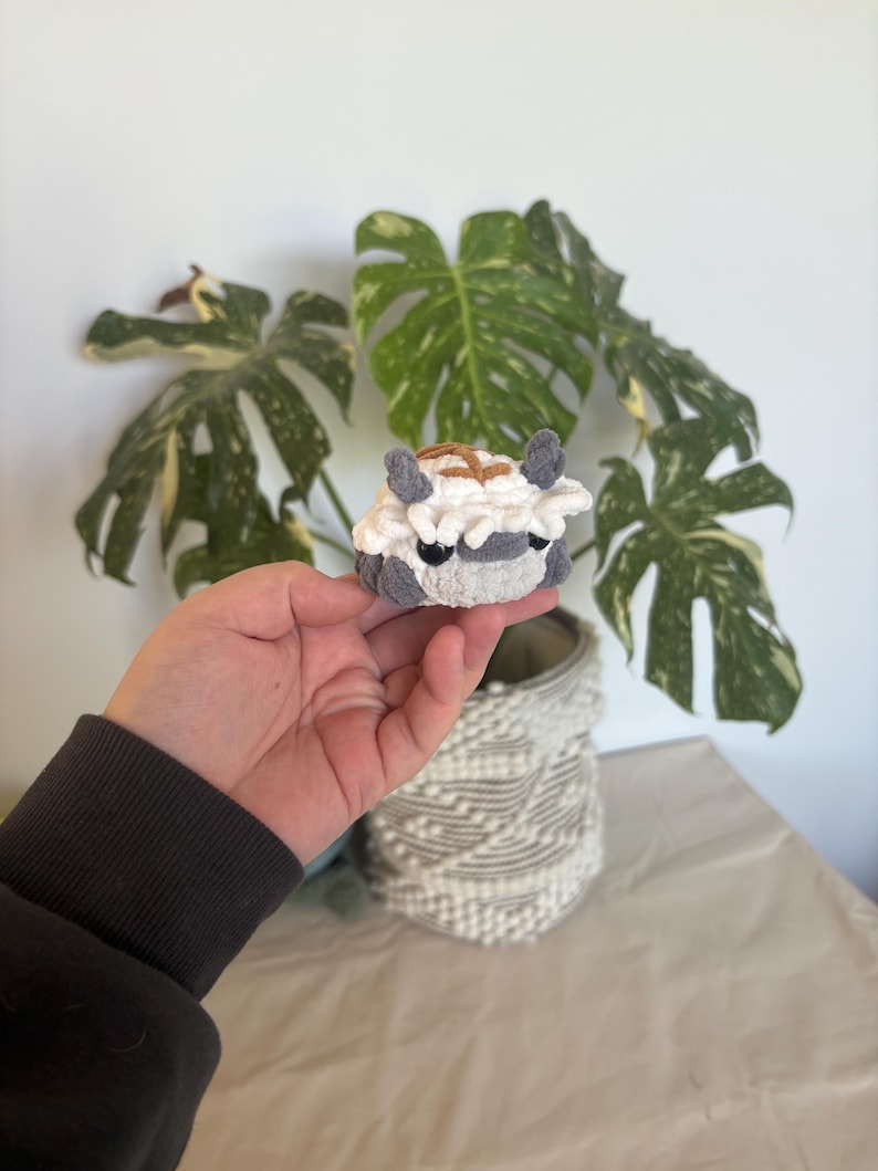 Op de afbeelding: Een kleine, handgemaakte gehaakte figuur van een wezen met grijze, witte en bruine details. De figuur heeft kleine oren en een rond lichaam. De figuur wordt vastgehouden voor een plant met grote groene bladeren met witte en gele vlekken.