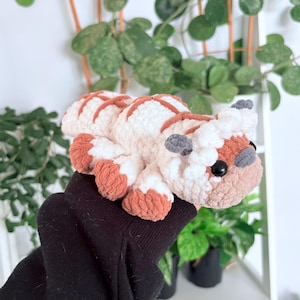 Puede incluir: Un peluche artesanal con forma de bisonte, con cuerpo blanco y rayas y detalles marrones. El bisonte tiene orejas grises, ojos negros y patas marrones. El peluche se sostiene en una manga negra.