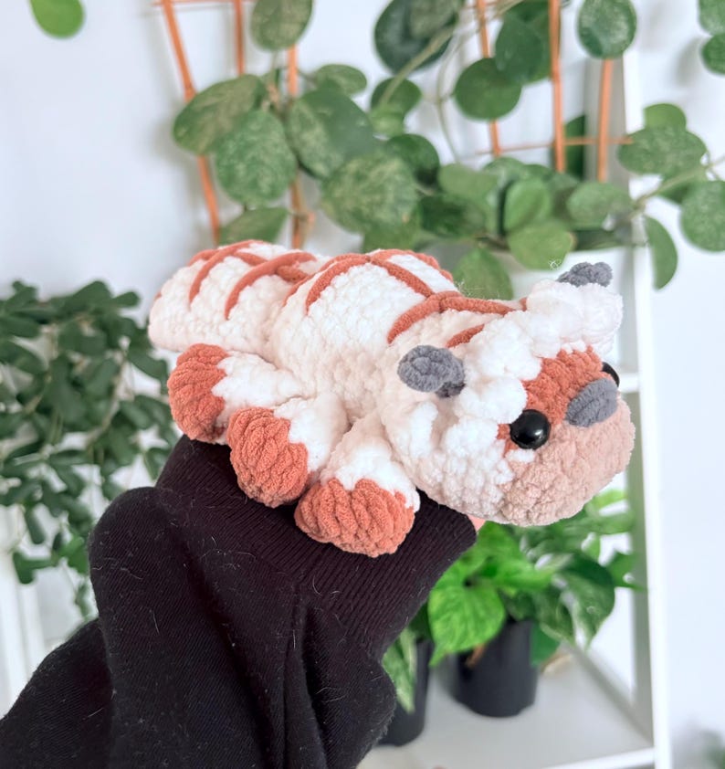 Puede incluir: Un peluche artesanal con forma de bisonte, con cuerpo blanco y rayas y detalles marrones. El bisonte tiene orejas grises, ojos negros y patas marrones. El peluche se sostiene en una manga negra.