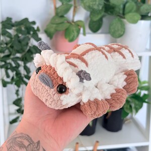 Puede incluir: Un peluche hecho a mano con ganchillo en forma de criatura blanca y marr&oacute;n con grandes ojos negros. El peluche tiene un aspecto suave y texturizado, con detalles marrones en los pies, la cabeza y la espalda. El juguete est&aacute; en la mano de una persona.