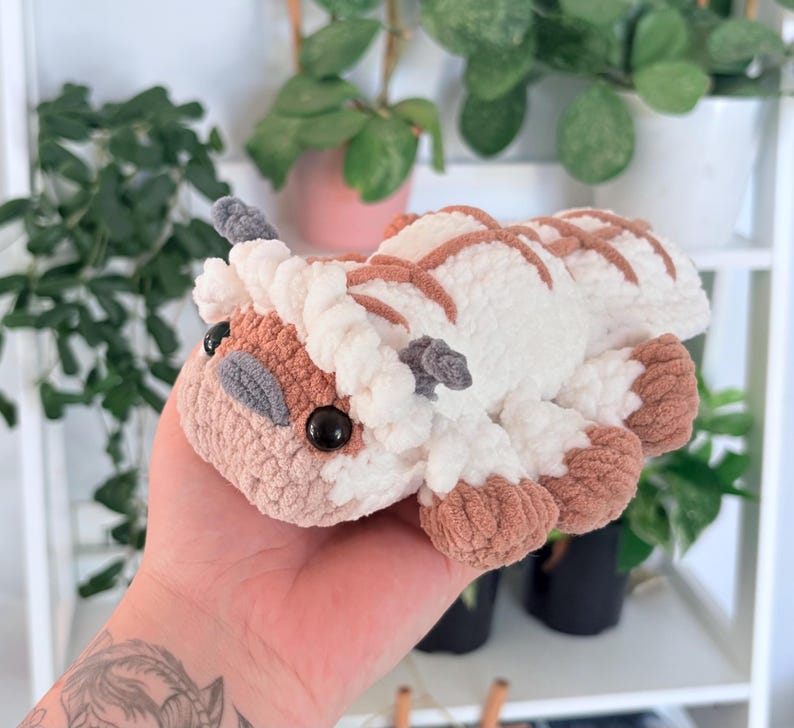 Puede incluir: Un peluche hecho a mano con ganchillo en forma de criatura blanca y marr&oacute;n con grandes ojos negros. El peluche tiene un aspecto suave y texturizado, con detalles marrones en los pies, la cabeza y la espalda. El juguete est&aacute; en la mano de una persona.