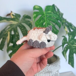 Op de afbeelding: Een kleine, handgemaakte gehaakte figuur van een bisonachtig wezen. Het pluche speelgoed is wit met bruine, grijze en zwarte accenten. De figuur heeft kleine grijze voetjes, oren en een staart. De achtergrond bevat groene bladeren.