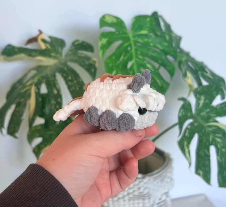 Op de afbeelding: Een kleine, handgemaakte gehaakte figuur van een bisonachtig wezen. Het pluche speelgoed is wit met bruine, grijze en zwarte accenten. De figuur heeft kleine grijze voetjes, oren en een staart. De achtergrond bevat groene bladeren.
