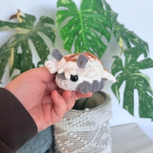 Op de afbeelding: Een klein, handgemaakt gehaakt dier in wit, bruin en grijs. Het pluche speelgoed heeft een rond lichaam met kleine, grijze aanhangsels en een paar kleine, witte vleugels. Een zwart oog is zichtbaar.