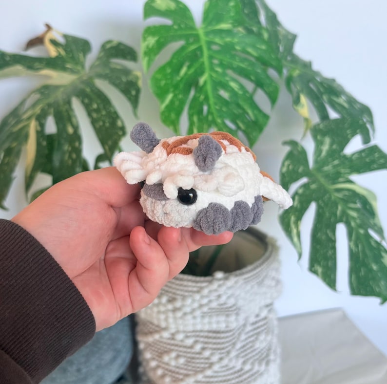 Op de afbeelding: Een klein, handgemaakt gehaakt dier in wit, bruin en grijs. Het pluche speelgoed heeft een rond lichaam met kleine, grijze aanhangsels en een paar kleine, witte vleugels. Een zwart oog is zichtbaar.