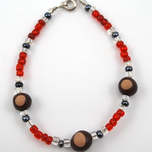 May include: A beaded bracelet featuring alternating red, clear, and dark blue beads. The bracelet includes three dark brown beads with a tan oval design. The bracelet is connected with a silver clasp.