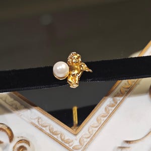 Pode incluir: Um colar gargantilha de veludo preto com um pingente de querubim dourado segurando uma pérola. O querubim é detalhado com asas e um rosto sorridente. O colar é exibido em um fundo decorativo branco e dourado.