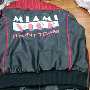 May include: Black jacket with red accents and the text "MIAMI VICE STUNT TEAM" on the back. The jacket has a ribbed black collar and cuffs, and a red collar. The jacket is made of a shiny material.