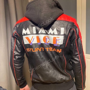 May include: A black leather jacket with red accents and a black hood. The back features the white text MIAMI, the orange and purple text VICE, and the orange text STUNT TEAM. The jacket has long sleeves, a ribbed hem, and red piping details.