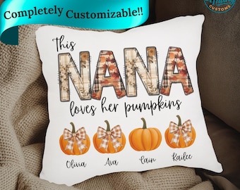 May include: White decorative pillow with the text "This NANA loves her pumpkins." The word "NANA" is in a fall-themed plaid pattern. Below are four orange pumpkins with names: Olivia, Ava, Cain, and Kailee.
