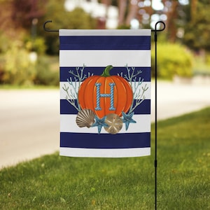 May include: A garden flag with a navy blue and white striped background. An orange pumpkin with a blue letter "H" is centered on the flag, surrounded by seashells, starfish, and coral. The flag is displayed outdoors on a black metal stand.