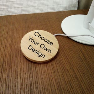 May include: A round, light-colored wooden coaster with the text "Choose Your Own Design" in black. The coaster is on a brown wooden surface, near a white lamp base.